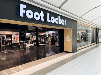 Entrada de Foot Locker en Willowbrook Mall, con letrero blanco, paneles de madera y exhibidores de tenis visibles desde el pasillo.