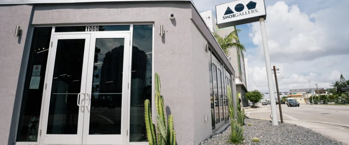 Fachada de Shoe Gallery en Miami, con paredes grises, una entrada de vidrio y cactus junto a la acera.