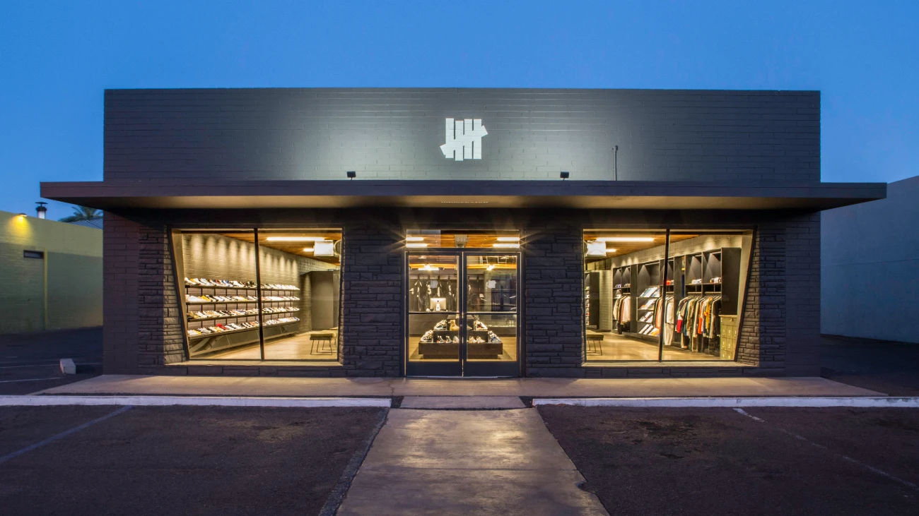 Undefeated storefront at dusk with illuminated sneaker displays behind large windows.