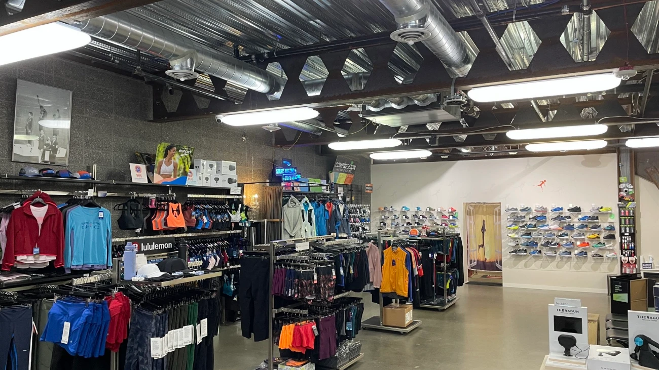Running shop interior with exposed ceiling ducts, apparel racks and a wall of shoes.
